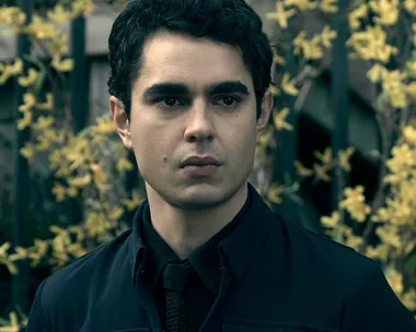 Nick from "The Handmaid's Tale" stands outdoors in front of yellow flowering plants, dressed in a dark coat, looking serious.