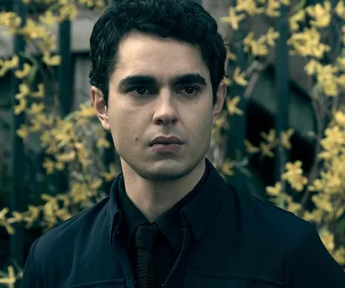 Nick from "The Handmaid's Tale" stands outdoors in front of yellow flowering plants, dressed in a dark coat, looking serious.