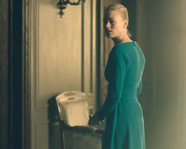 Serena Joy from "The Handmaid's Tale" in a green dress, standing in a dimly lit, ornate room, looking over her shoulder.