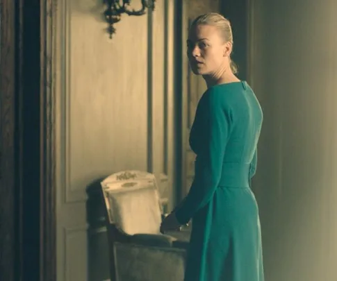 Serena Joy from "The Handmaid's Tale" in a green dress, standing in a dimly lit, ornate room, looking over her shoulder.