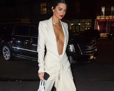 A woman wearing an off-white plunging outfit holds a tiny white handbag and a phone, walking outside at night.