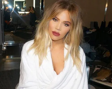 Khloe Kardashian sitting in makeup chair with blonde hair and white robe, posing for a photo.