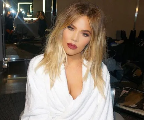 Khloe Kardashian sitting in makeup chair with blonde hair and white robe, posing for a photo.