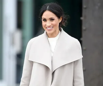 Meghan Markle smiles wearing a light-gray coat over a white top.