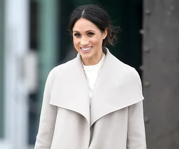 Meghan Markle smiles wearing a light-gray coat over a white top.