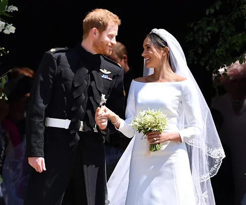 Bride and groom holding hands and smiling at each other outside, bride in white dress and veil, groom in military uniform.