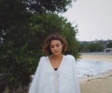 Woman in white sweater stands near a tree with a beach and water in the background.