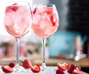 Two glasses of pink gin with ice and sliced strawberries, surrounded by fresh strawberries on a table.