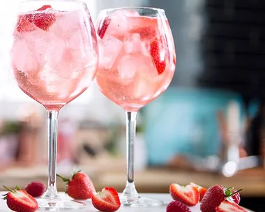 Two glasses of pink gin with ice and sliced strawberries, surrounded by fresh strawberries on a table.