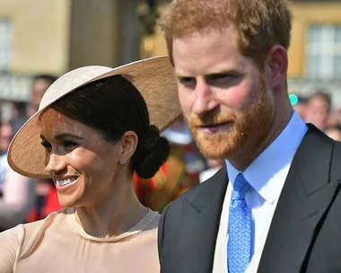 Prince Harry and Meghan Markle smiling while attending an outdoor event, with Harry in a suit and Meghan in a light hat.