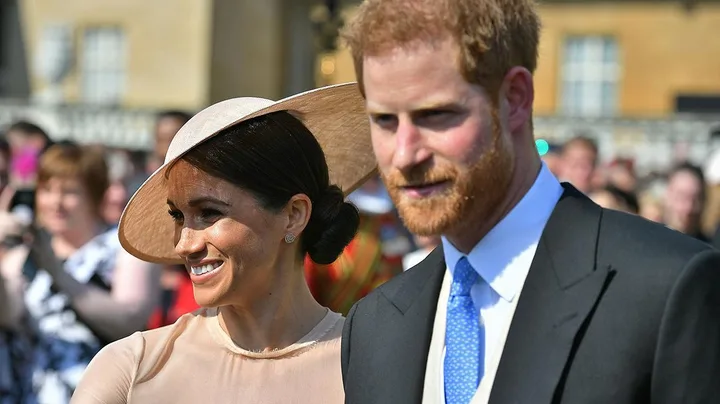 Prince Harry and Meghan Markle smiling while attending an outdoor event, with Harry in a suit and Meghan in a light hat.