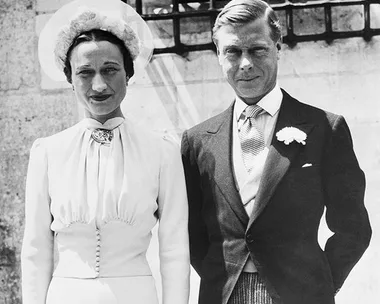 Black-and-white photo of Wallis Simpson in a light-colored dress and hat, standing beside Edward VIII in a suit with a flower boutonniere.