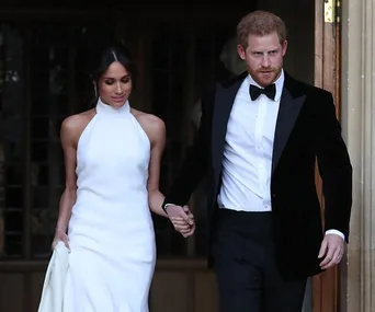 Meghan Markle in a white Stella McCartney reception dress with Prince Harry, holding hands while exiting a building.