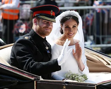 Prince Harry in military uniform and Meghan Markle in wedding dress wave while riding in an open carriage.