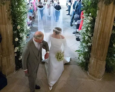 The bride walks down the aisle with an older man in a suit at a wedding ceremony, followed by flower girls and boys.