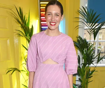 A woman in a pink dress stands in front of a yellow wall with plants and a neon light behind her.