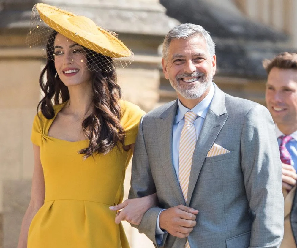 Two elegantly dressed individuals, woman in yellow dress and hat, man in gray suit, smile arm-in-arm, outdoors at royal wedding.