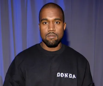 Kanye West wearing a black "DONDA" sweatshirt, standing against a backdrop of blue and grey curtains.