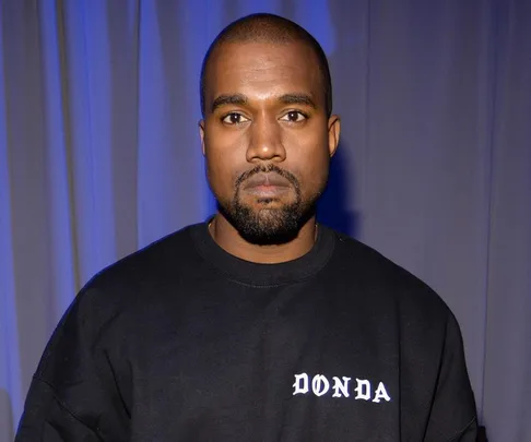 Kanye West wearing a black "DONDA" sweatshirt, standing against a backdrop of blue and grey curtains.