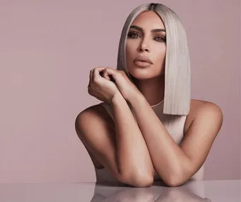 Kim Kardashian with straight platinum blonde hair and nude makeup, resting her arms on a reflective surface against a beige background.