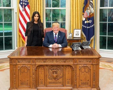 Kim Kardashian standing next to Donald Trump seated at the Resolute Desk in the Oval Office with flags in the background.