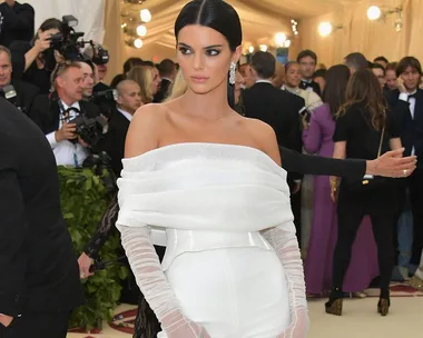 A woman in an off-shoulder white dress poses on the red carpet at an event, surrounded by photographers and attendees.