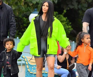 A woman in a neon green jacket holding hands with two children, one in an orange top and the other in black, at Disneyland.