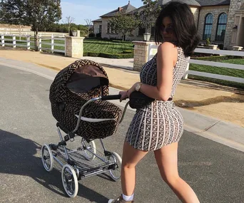 A woman in a patterned dress pushes a matching patterned baby stroller on a road near a large house.