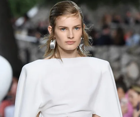 Model in a white outfit with large silver earrings, walking the runway at the Louis Vuitton Cruise 2019 show.