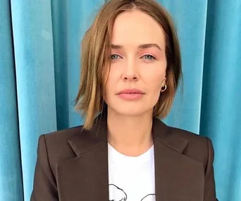 A woman with short brown hair wearing a brown blazer and white shirt stands in front of a blue curtain.