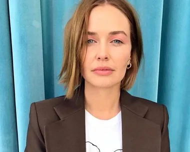 A woman with short brown hair wearing a brown blazer and white shirt stands in front of a blue curtain.