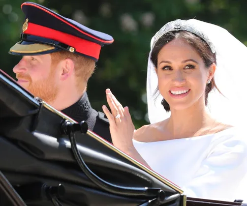 Prince Harry in uniform and Meghan Markle in wedding dress smile and wave from a carriage at their royal wedding.