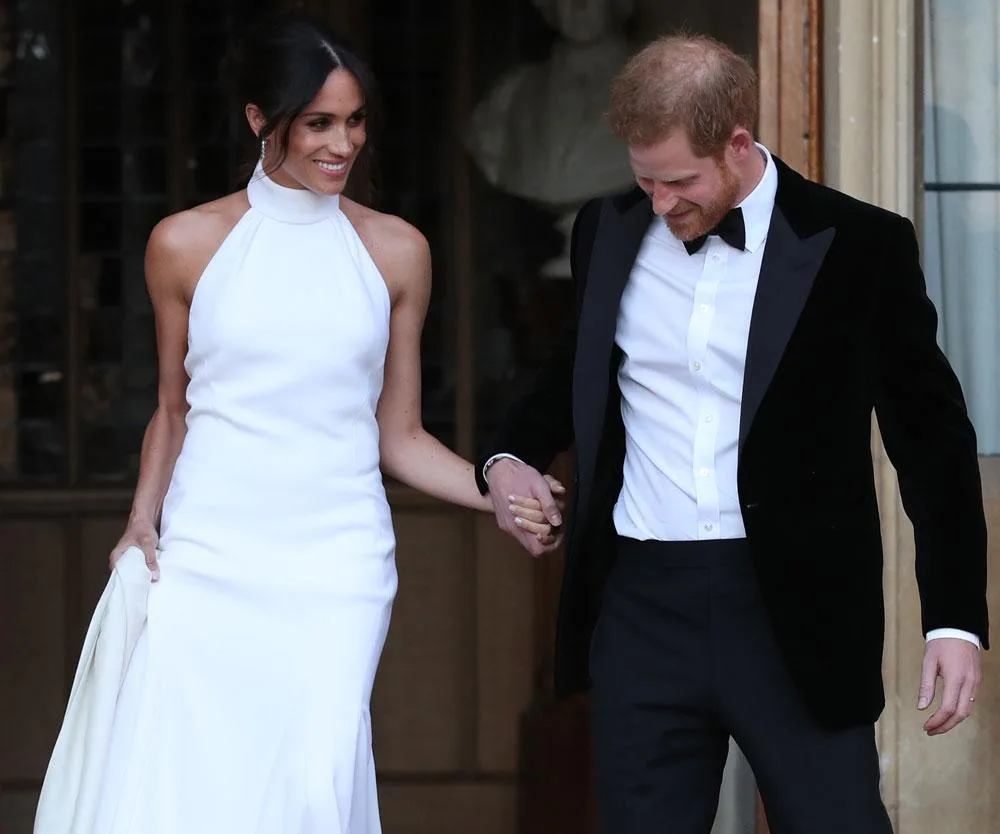 A man in a tuxedo and a woman in a white halter dress hold hands and smile, standing outside a building.