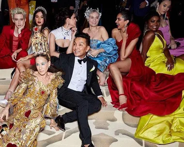 A group of celebrities in colorful, extravagant dresses at the Met Gala 2018, sitting together on the carpet.