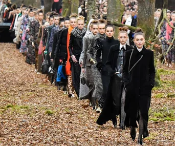 Models walking in a fashion show outdoors, wearing fall and winter outfits, with trees and leaves around.