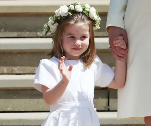 Young girl in a white dress and floral crown holding hands and waving on wedding steps.