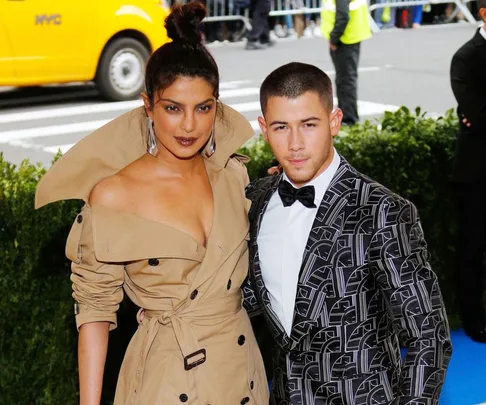 Priyanka Chopra in a beige trench coat and Nick Jonas in a patterned tuxedo at an outdoor event with a yellow NYC taxi in the background.