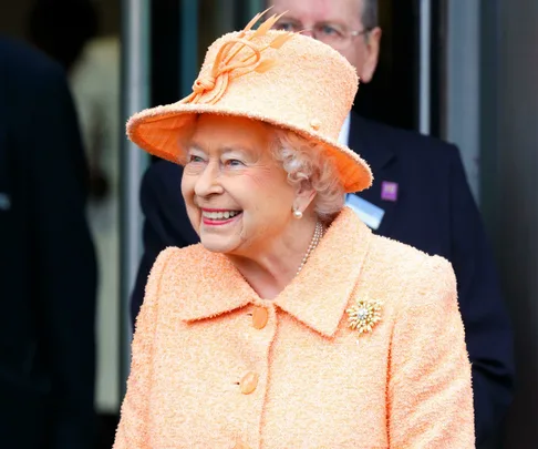 Elderly woman in a peach ensemble, including a textured coat, matching hat, pearl earrings, and brooch, smiles warmly.