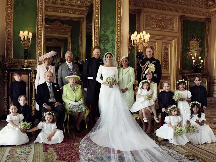 Royal wedding family portrait with the bride and groom at center, surrounded by family members and flower girls in a regal room.