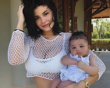 A woman with short dark hair in a white mesh top holds a baby in a white outfit outdoors.