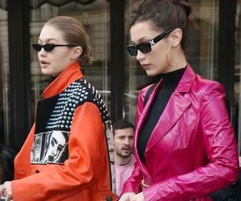 Two women in stylish outfits and sunglasses walk outdoors. One in orange jacket with studs and print, other in pink blazer.