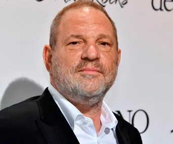 Close-up of a man with short hair and a beard, wearing a black suit and white shirt, standing against a white background with text.