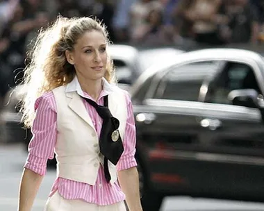 Carrie Bradshaw, in pink and white outfit with black tie, walking on city street in 'Sex and the City'.