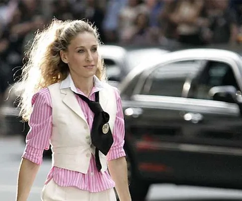 Carrie Bradshaw, in pink and white outfit with black tie, walking on city street in 'Sex and the City'.