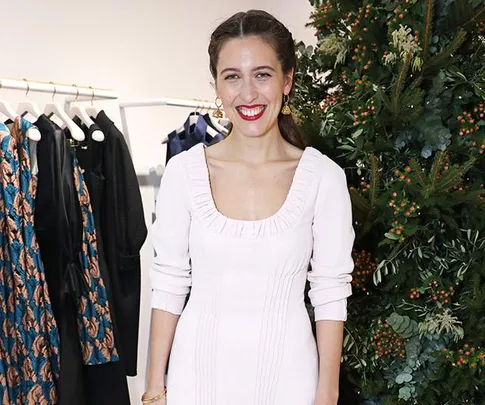 A woman in a white dress stands smiling in front of a clothing rack and a decorated plant.