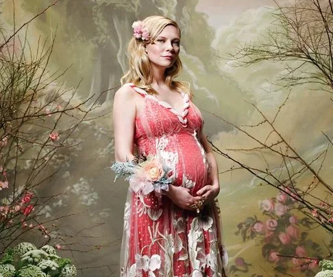 Pregnant woman in a floral dress posing in front of a whimsical, nature-themed backdrop.