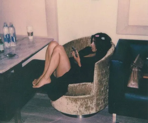 A woman in a robe with hair clips relaxes in a chair, looking at her phone, preparing for the Met Gala with bottled water nearby.