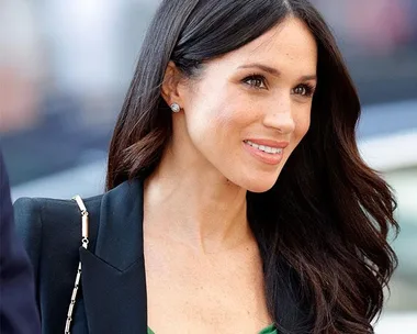 Meghan Markle smiling, wearing a green dress and black blazer, with long brown hair down.