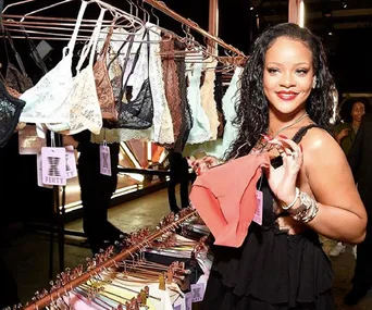 Rihanna displays Fenty lingerie in a store, holding a coral-colored piece, smiling, surrounded by various intimate apparel.