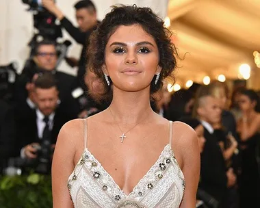 A woman with dark, curly hair wearing a white dress and a cross necklace at a formal event, posing for photos with people in the background.